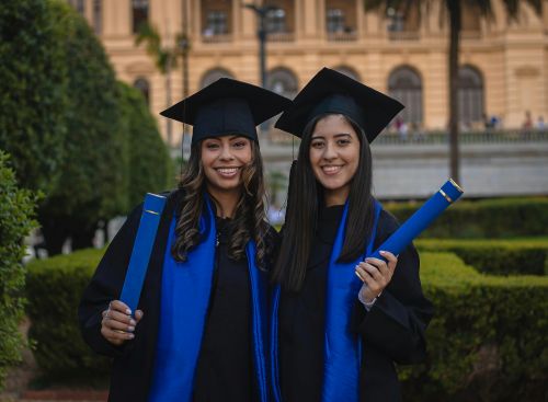 Two young women wearing graduation caps and gowns smile while holding diplomas